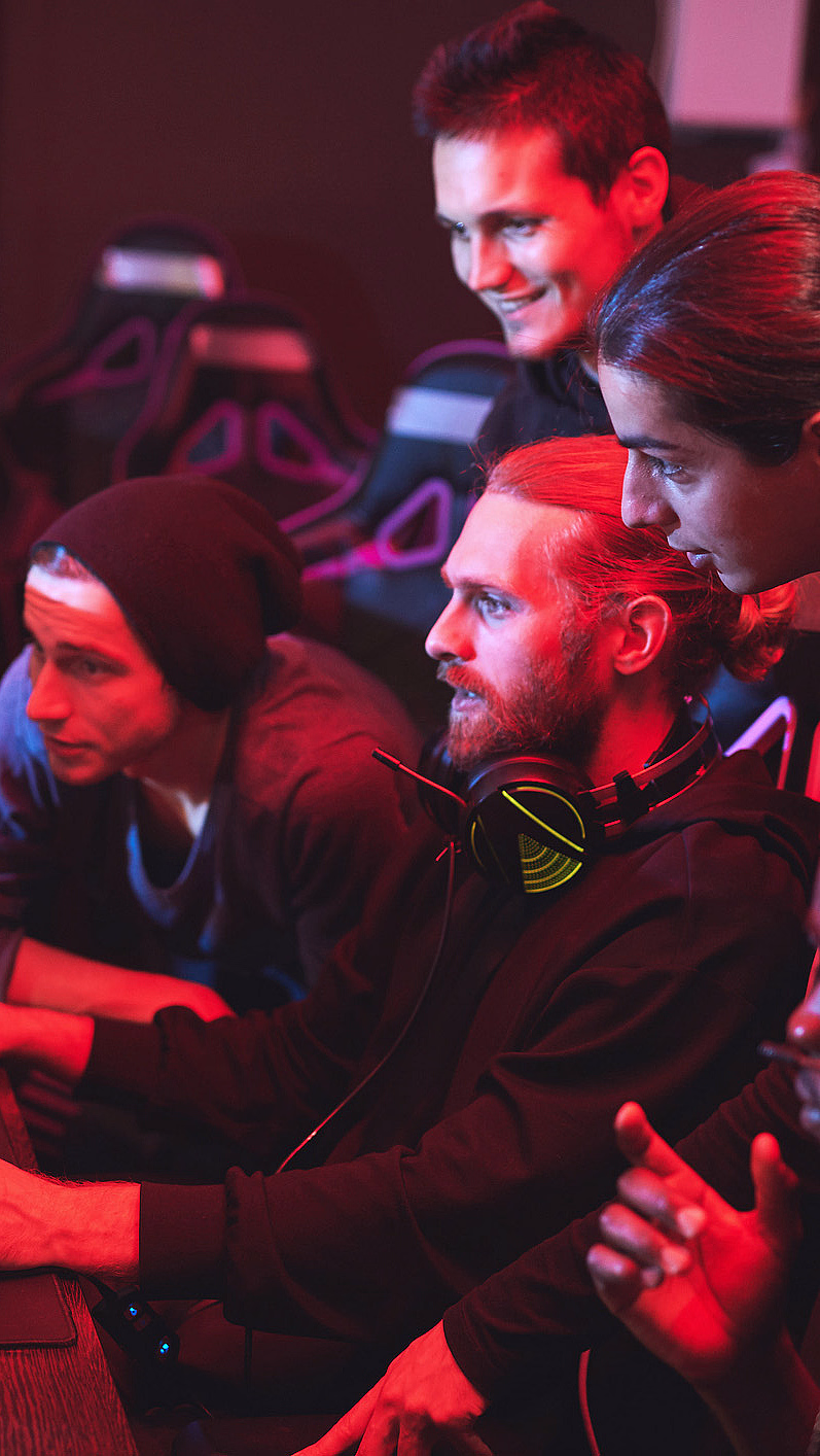 Fire personer med headset ser på en dataskjerm i et rom med rødt lys og gamingstoler