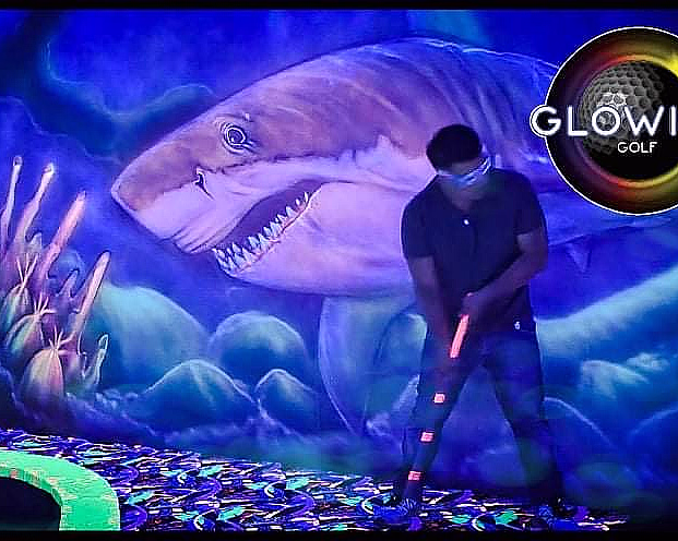 Person spiller minigolf i neonfarger med en lysende hai på veggen og logoen Glowing Golf