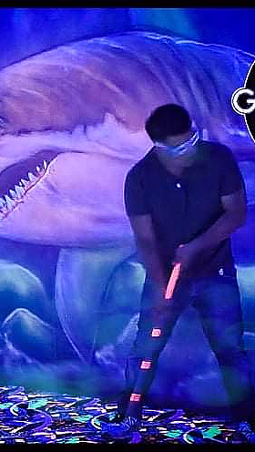 Person spiller minigolf i neonfarger med en lysende hai på veggen og logoen Glowing Golf