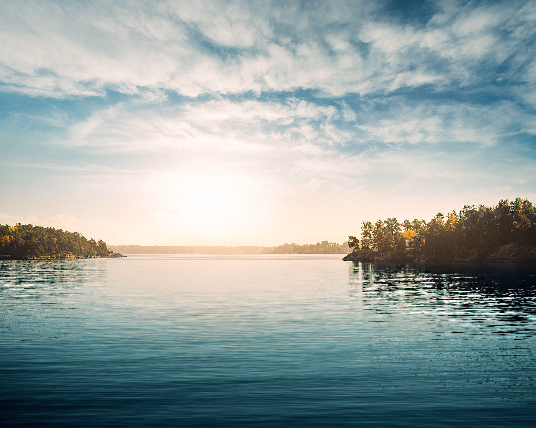 Blikkstille vann som reflekterer blå himmel, hvite skyer og lav sol ved en skogkledd horisont