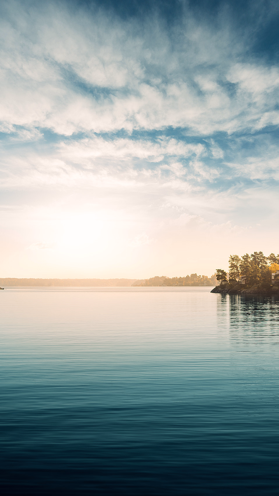 Blikkstille vann som reflekterer blå himmel, hvite skyer og lav sol ved en skogkledd horisont