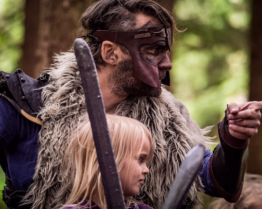 Mann i vikingkostyme med skinnmaske og pels som peker fremover ved siden av et barn