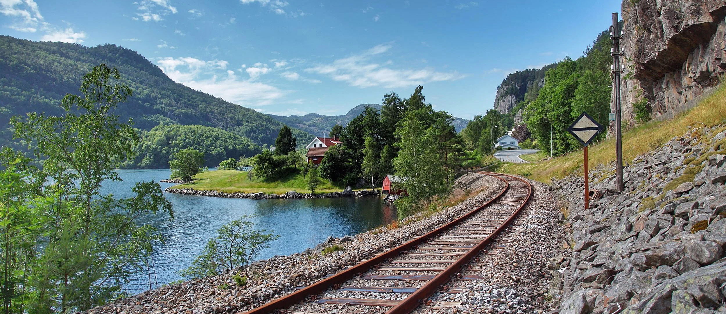 Jernbanespor langs en blå innsjø med et lite rødt hus og fjell i bakgrunnen