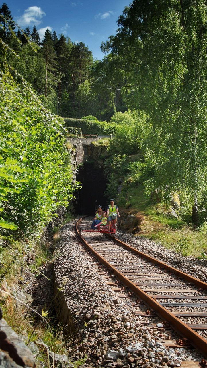 To personer i sikkerhetsvester på dresin på skinner gjennom en grønn skog mot en tunnel