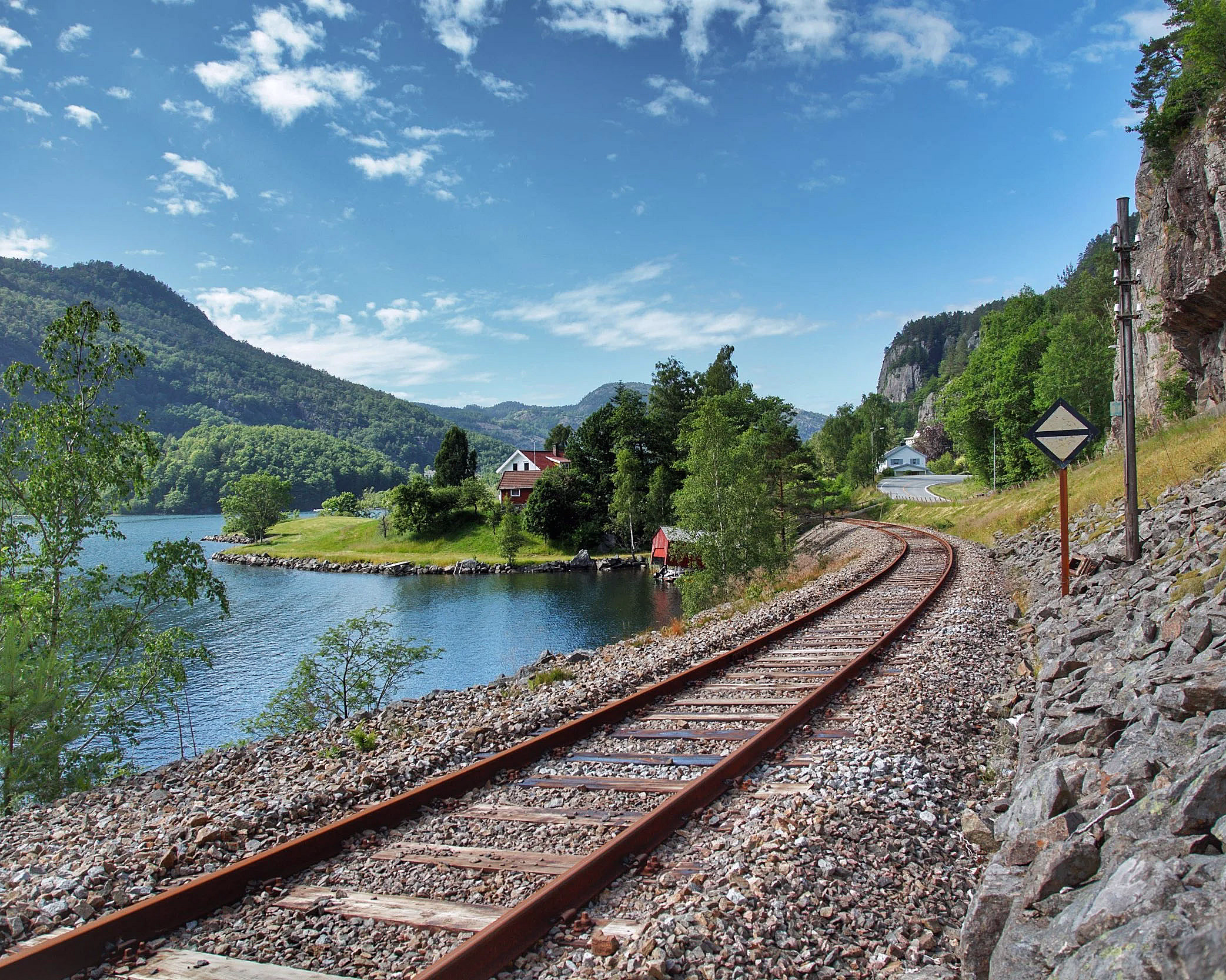 Svingete jernbanespor langs en blå innsjø med et lite rødt hus og grønne åser