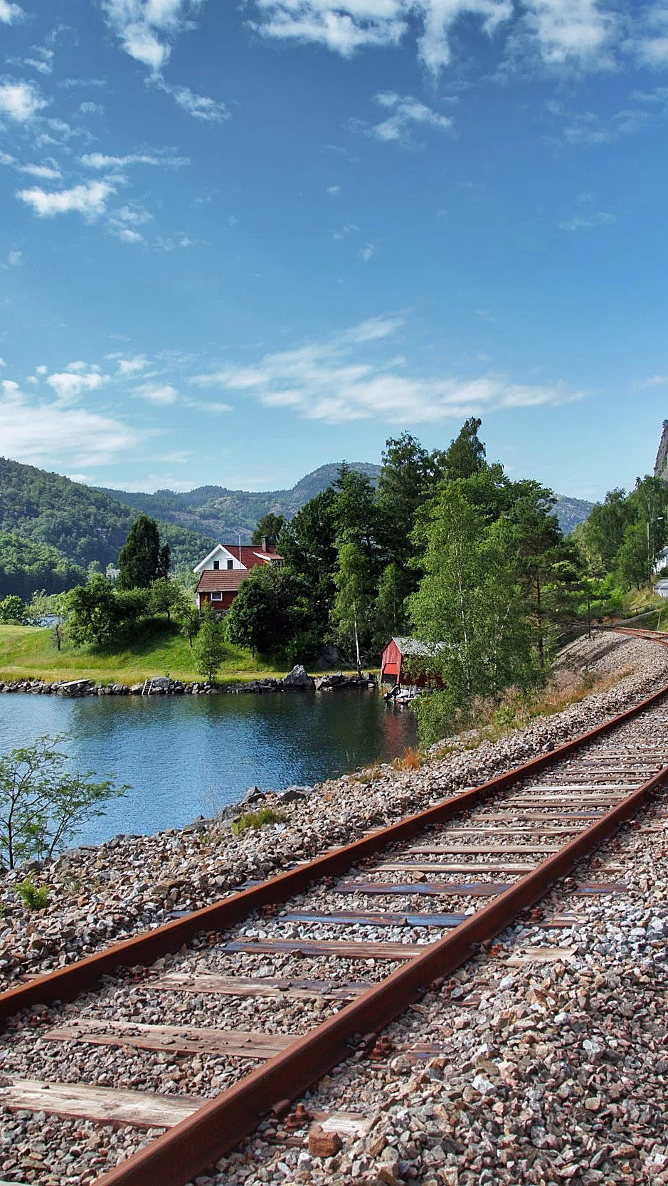 Svingete jernbanespor langs en blå innsjø med et lite rødt hus og grønne åser
