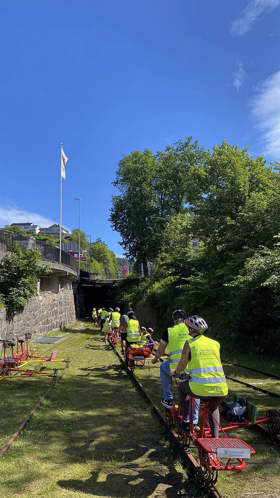 Personer i gule vester og hjelmer sykler på røde dresiner langs en jernbanelinje