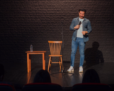 Mann med mikrofon på en scene foran en mørk murvegg og et publikum