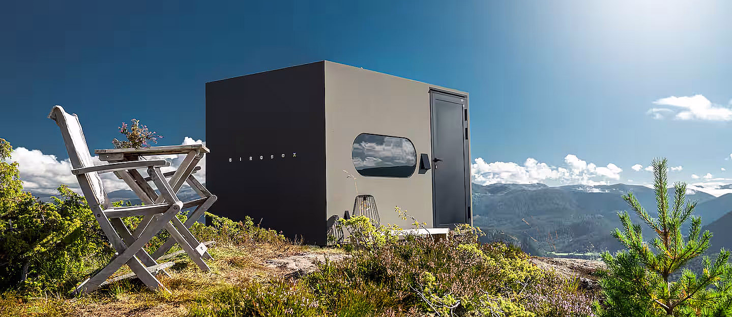 En moderne, liten svart hytte på en gresskledd bakketopp med utemøbler og utsikt over fjellene under en klar blå himmel.
