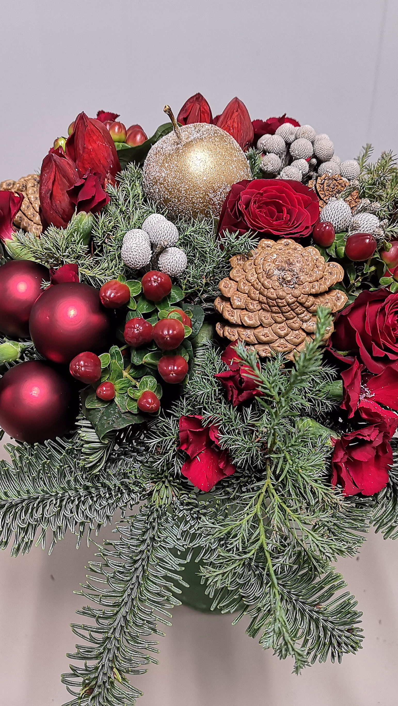 Festlig juleoppsats med røde roser, amaryllis, grønt bar, kongler og skinnende julekuler i rødt og gull.