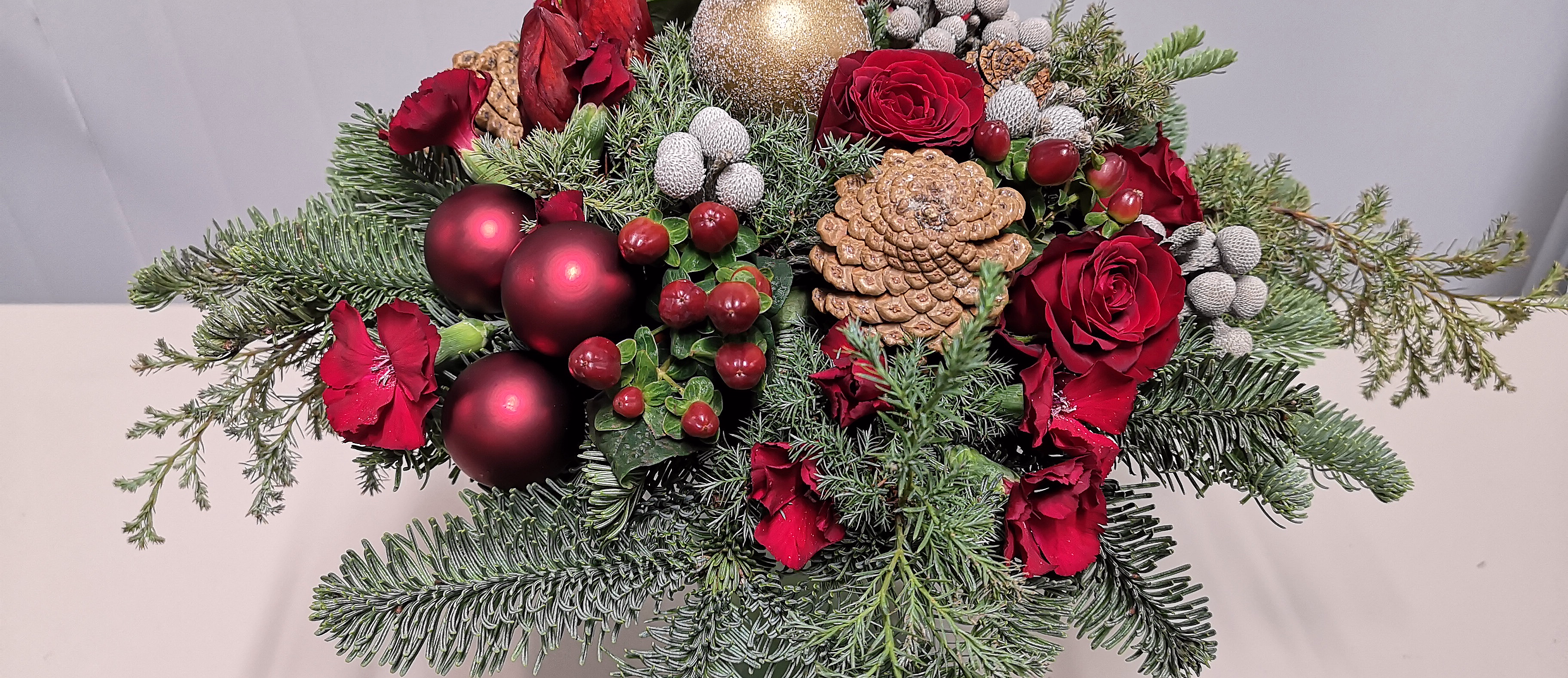 Festlig juleoppsats med røde roser, amaryllis, grønt bar, kongler og skinnende julekuler i rødt og gull.