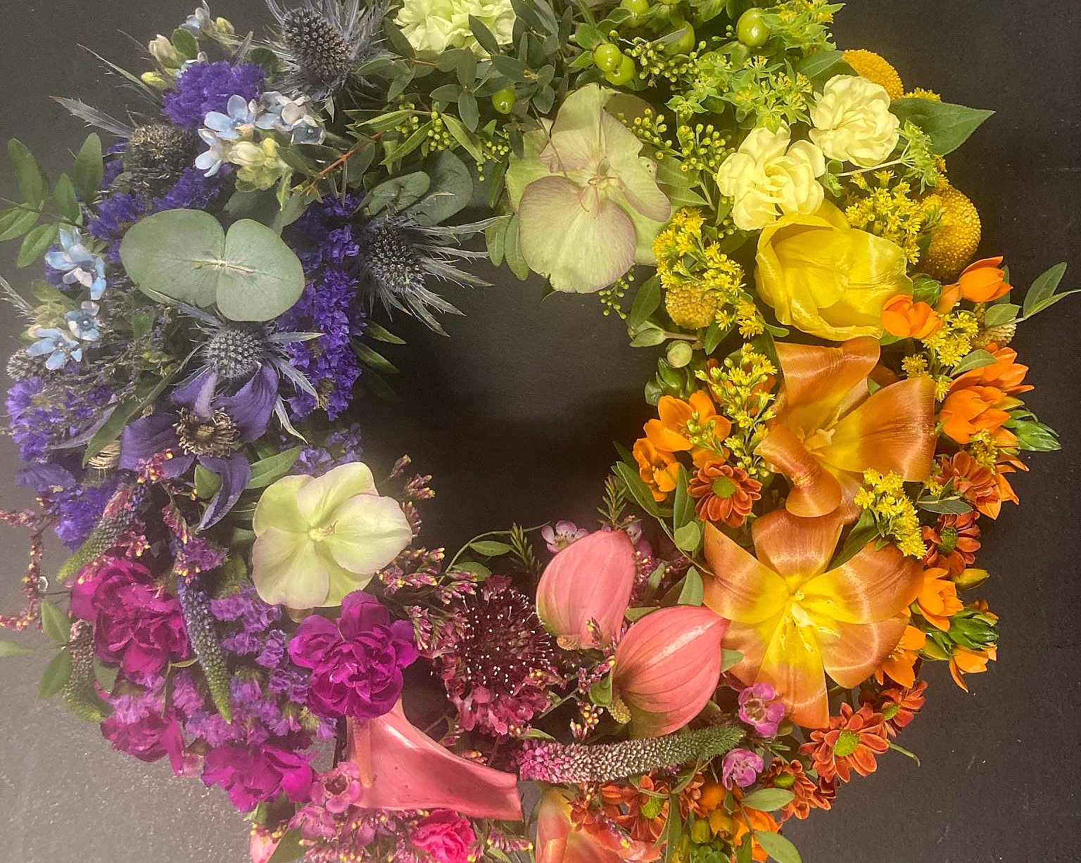 En sirkulær blomsterkrans med en fargerik overgang fra lilla og blått til grønt, gult, oransje og rosa, bestående av roser, liljer og tistler på en mørk bakgrunn.