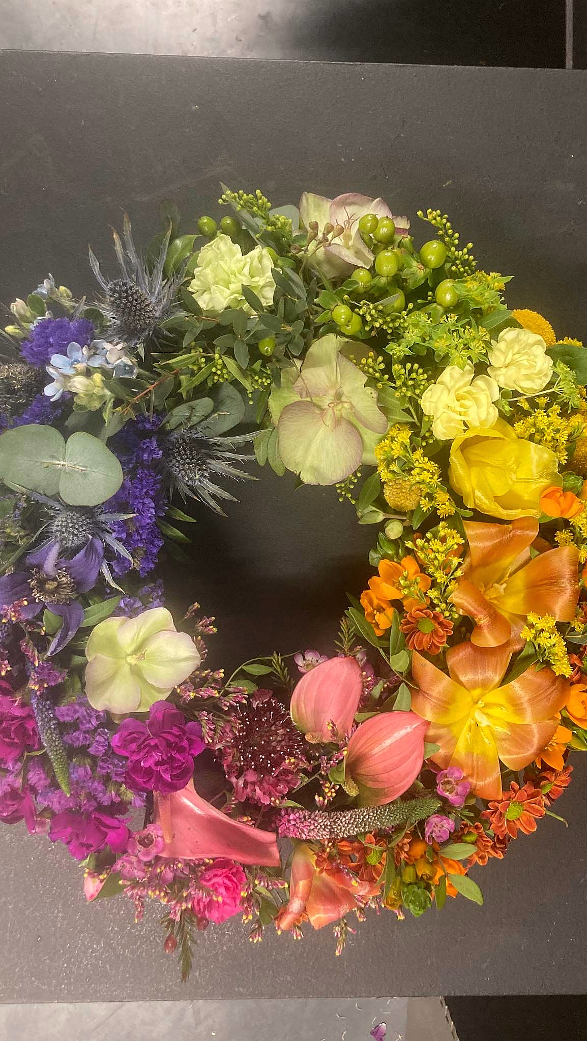 En sirkulær blomsterkrans med en fargerik overgang fra lilla og blått til grønt, gult, oransje og rosa, bestående av roser, liljer og tistler på en mørk bakgrunn.