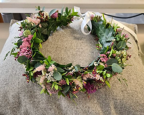 En sirkelformet blomsterkrans med grønne blader og blomster i rosa, lilla og hvitt, liggende på en grå, teksturert pute med et hvitt bånd.