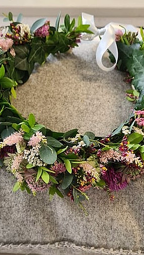 En sirkelformet blomsterkrans med grønne blader og blomster i rosa, lilla og hvitt, liggende på en grå, teksturert pute med et hvitt bånd.