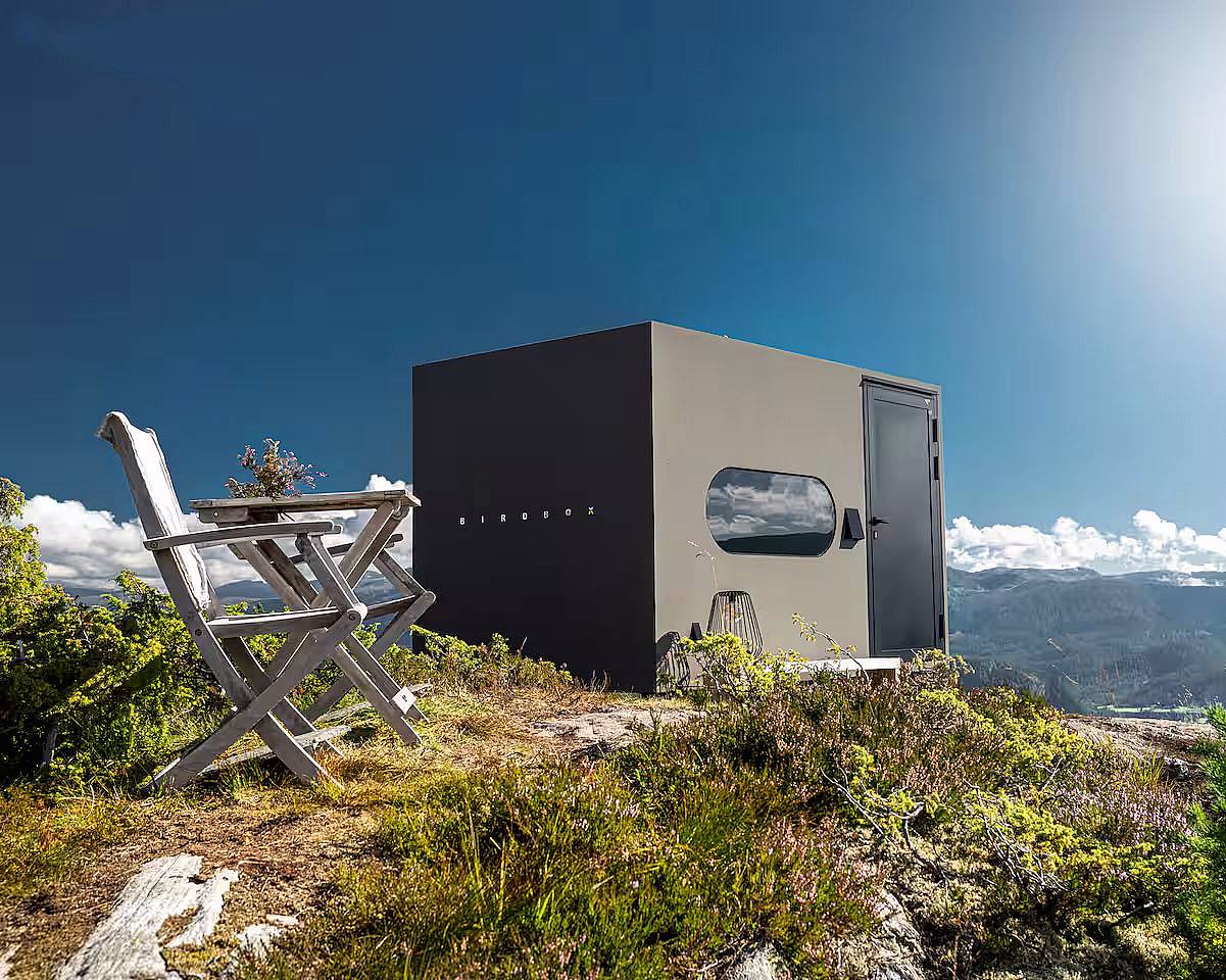 En liten, moderne svart hytte på en gresskledd høyde med utemøbler og utsikt over fjell under en klar blå himmel.