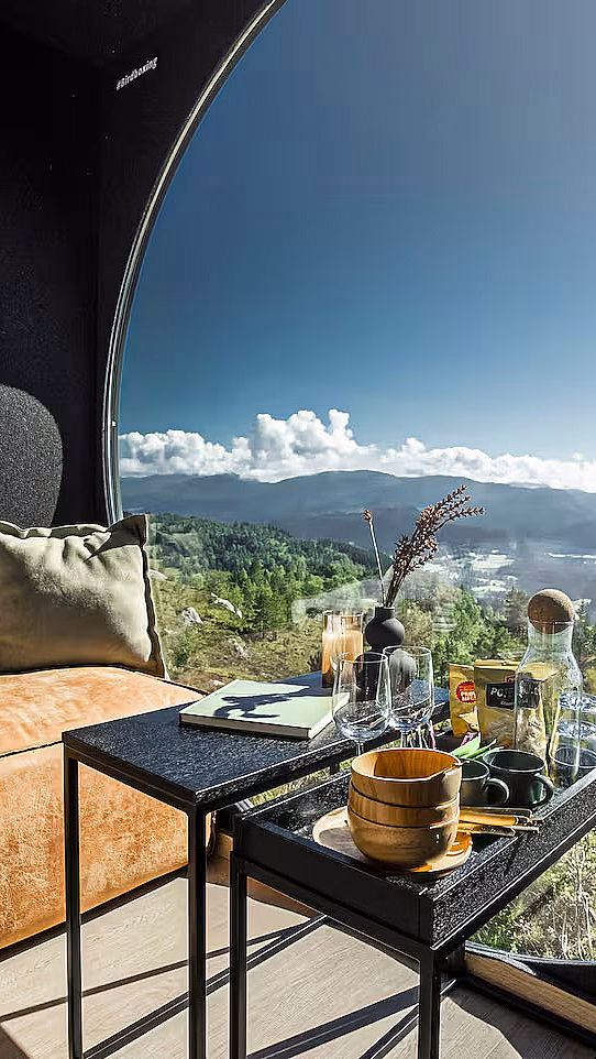 Et koselig interiør med et stort sirkulært vindu som viser utsikt over grønne fjell. En oransje sofa med en pute og et teppe står ved siden av to små svarte bord med en karaffel og kopper.
