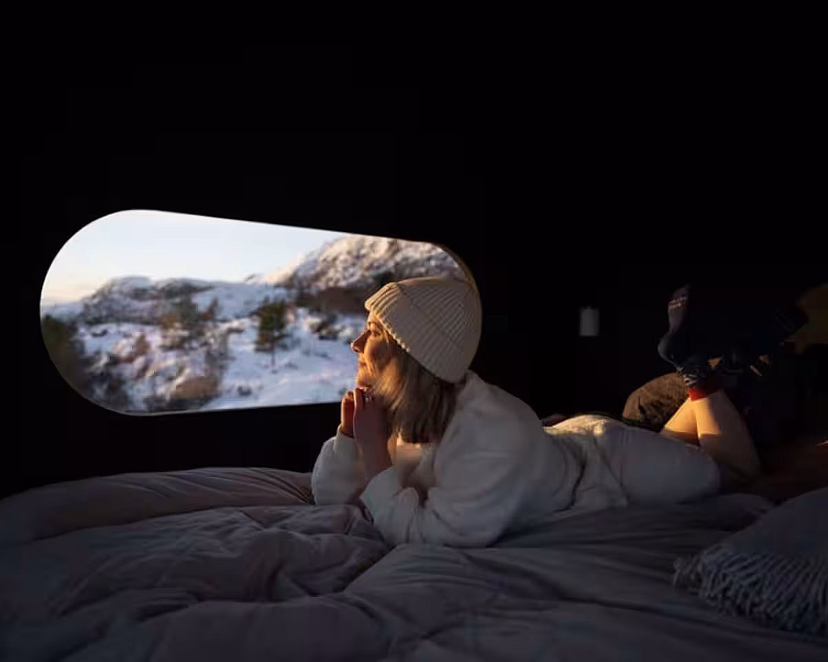 En person i hvit genser og lue ligger på en seng og ser ut på et snødekt landskap gjennom et ovalt vindu.