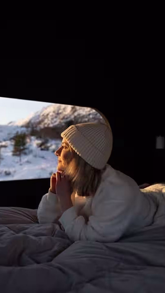 En person i hvit genser og lue ligger på en seng og ser ut på et snødekt landskap gjennom et ovalt vindu.