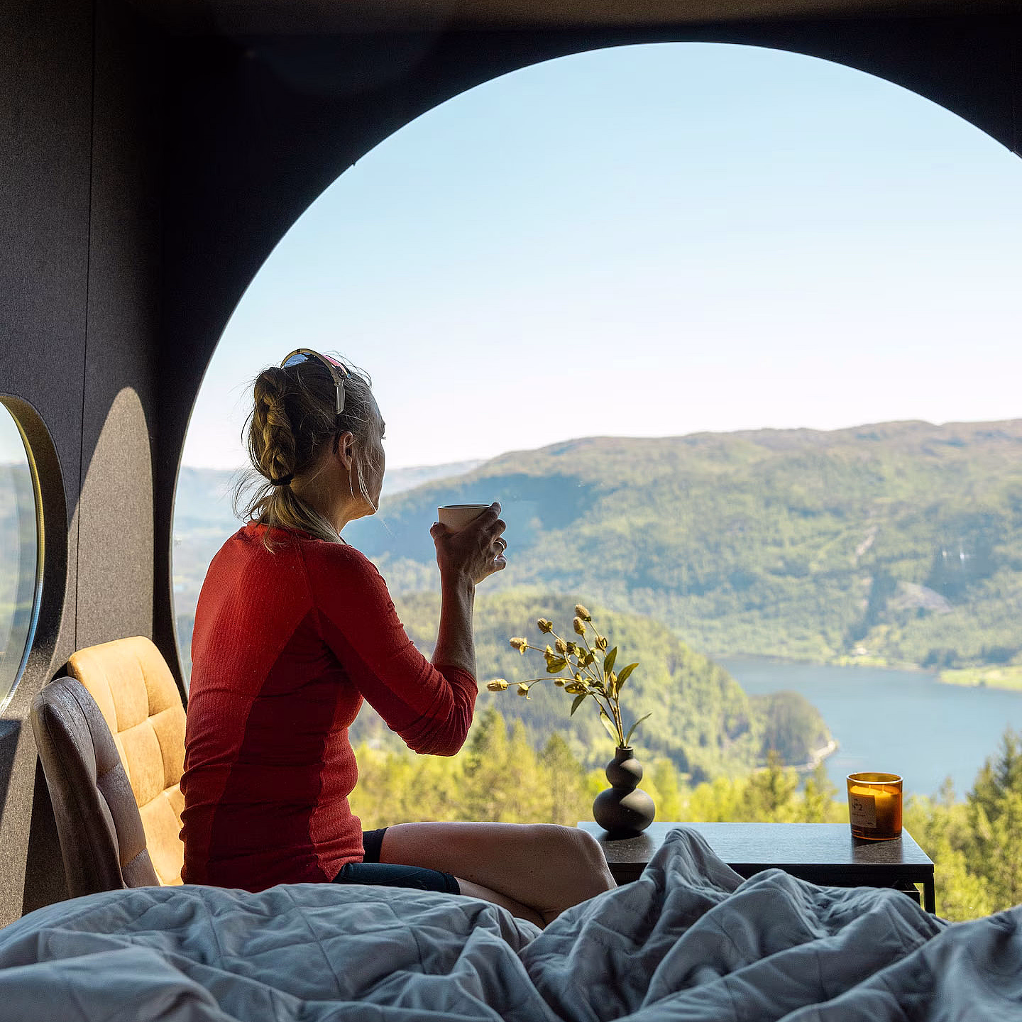 En person i rød genser sitter på en seng og ser ut gjennom et stort, rundt vindu mot grønne åser og en innsjø. Ved siden av sengen står et lite bord med gule blomster og et tent lys.
