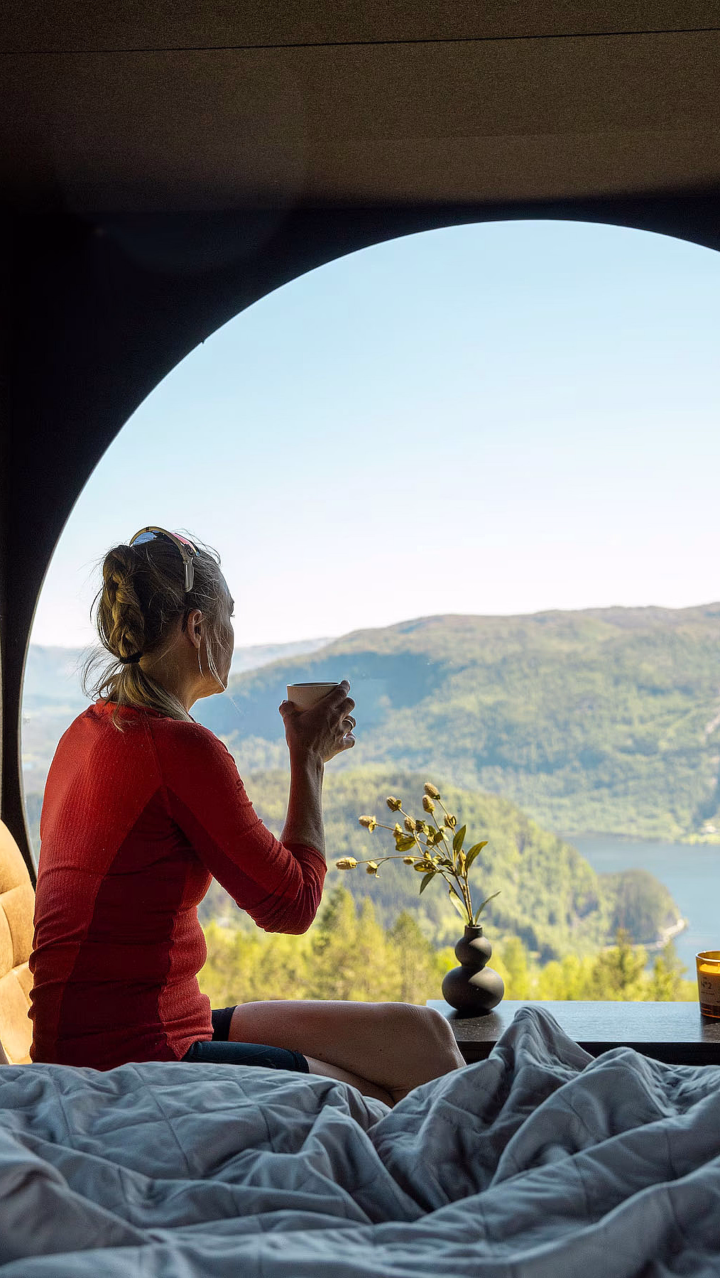 En person i rød genser sitter på en seng og ser ut gjennom et stort, rundt vindu mot grønne åser og en innsjø. Ved siden av sengen står et lite bord med gule blomster og et tent lys.
