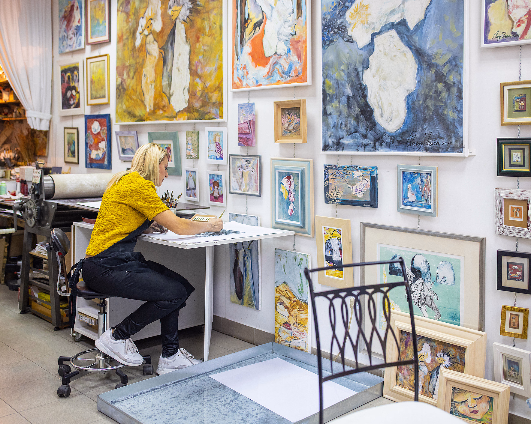 En person med lyst hår og gul skjorte sitter ved et skrivebord og tegner i et lyst kunststudio dekorert med fargerike malerier, med en trykkpresse og kunstutstyr i bakgrunnen.