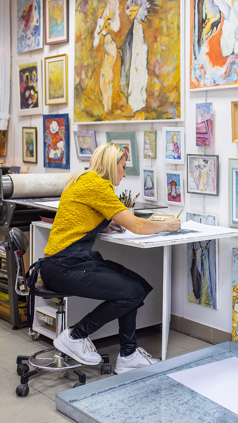 En person med lyst hår og gul skjorte sitter ved et skrivebord og tegner i et lyst kunststudio dekorert med fargerike malerier, med en trykkpresse og kunstutstyr i bakgrunnen.