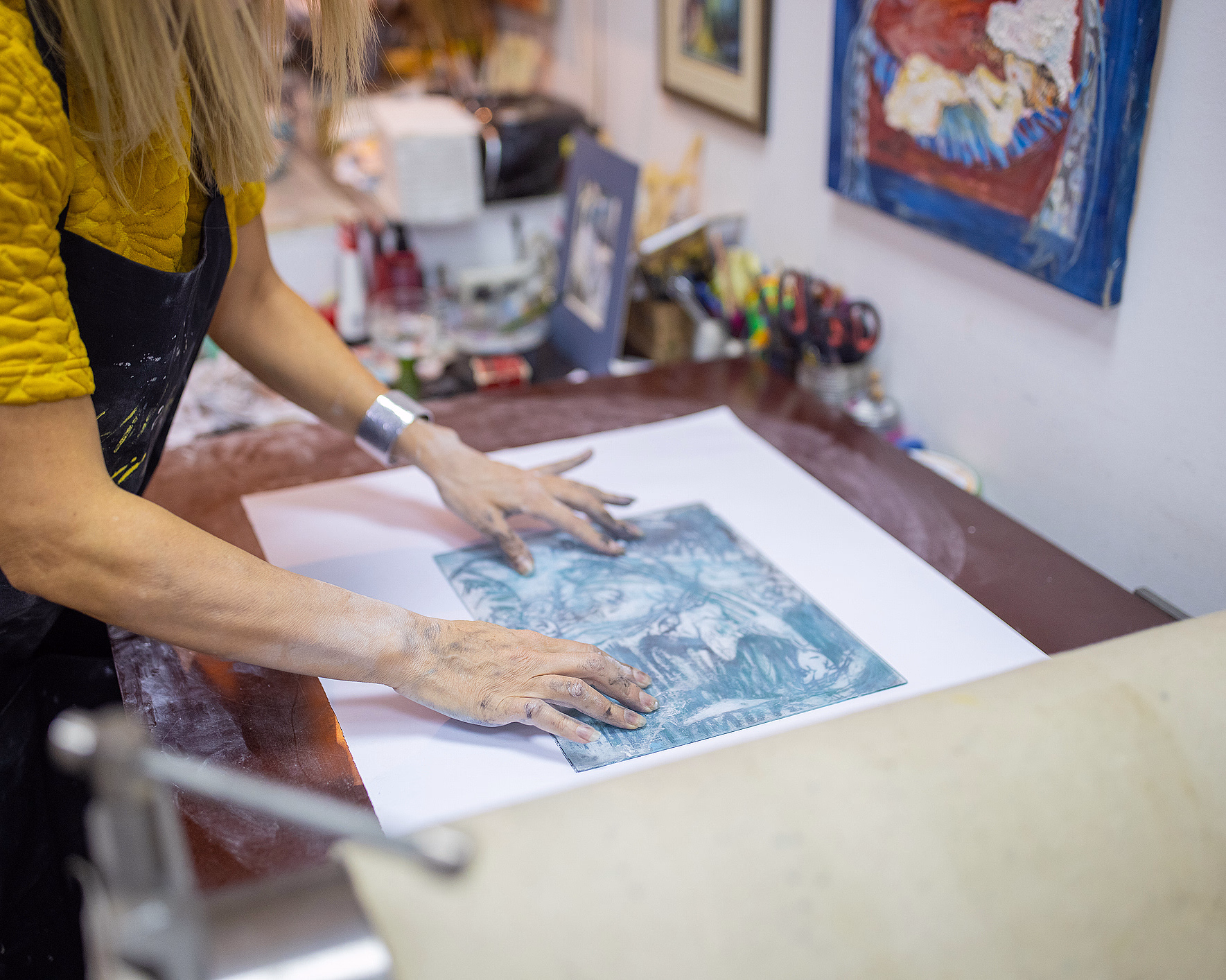 En kunstner med svertete hender i gul skjorte og mørkt forkle legger et blått og hvitt grafisk trykk på et papir i et lyst atelier.