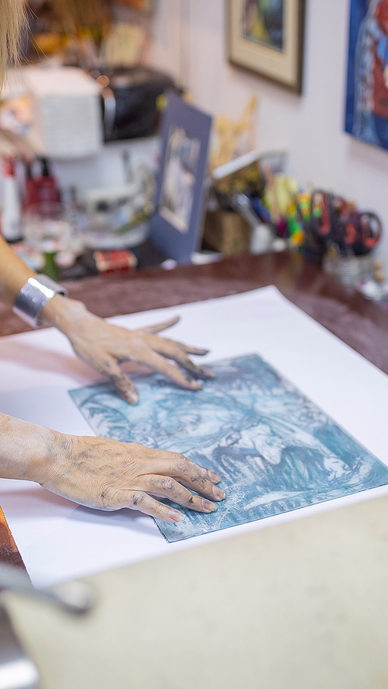 En kunstner med svertete hender i gul skjorte og mørkt forkle legger et blått og hvitt grafisk trykk på et papir i et lyst atelier.