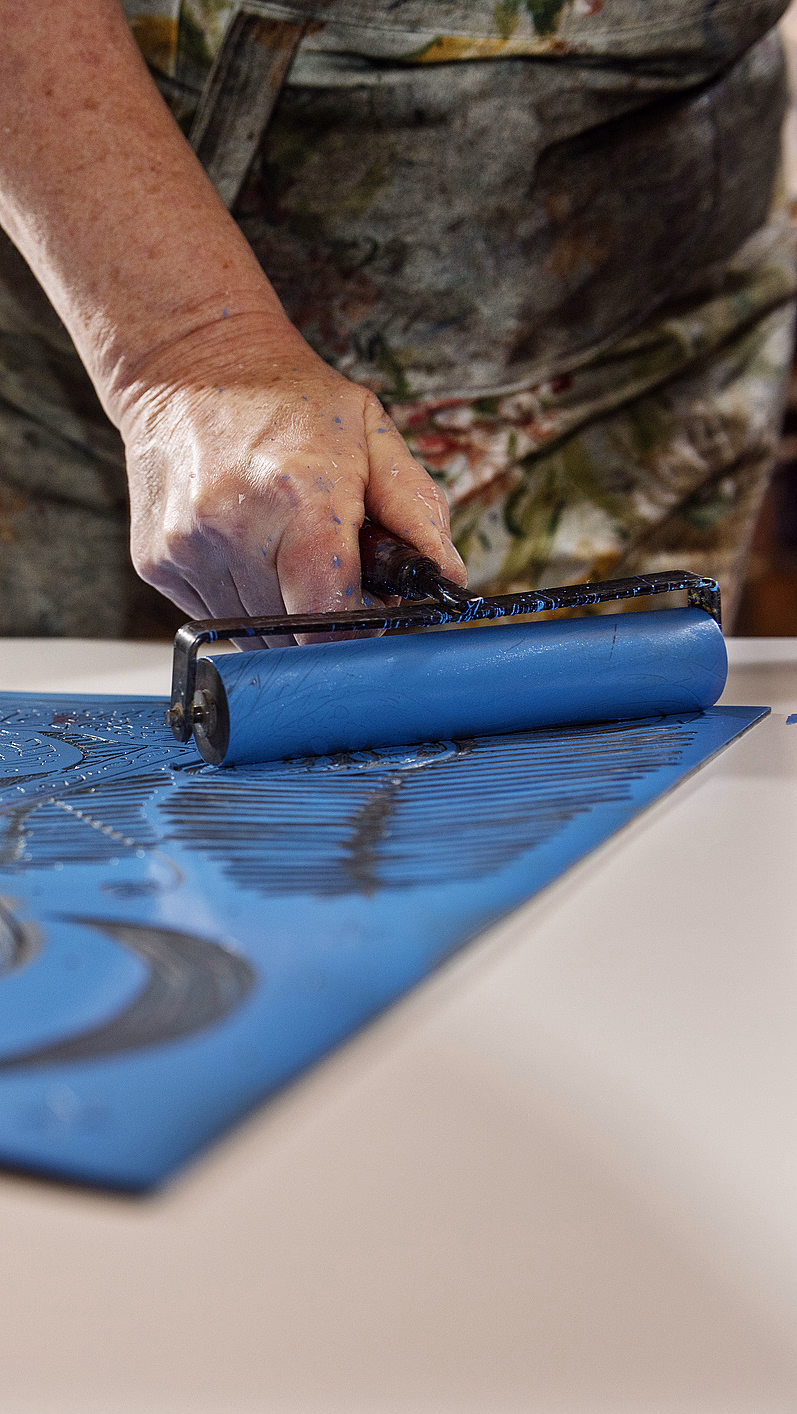 En person som påfører blått blekk på en utskåret linoleumplate med bladmønster ved bruk av en rulle i et kunststudio.