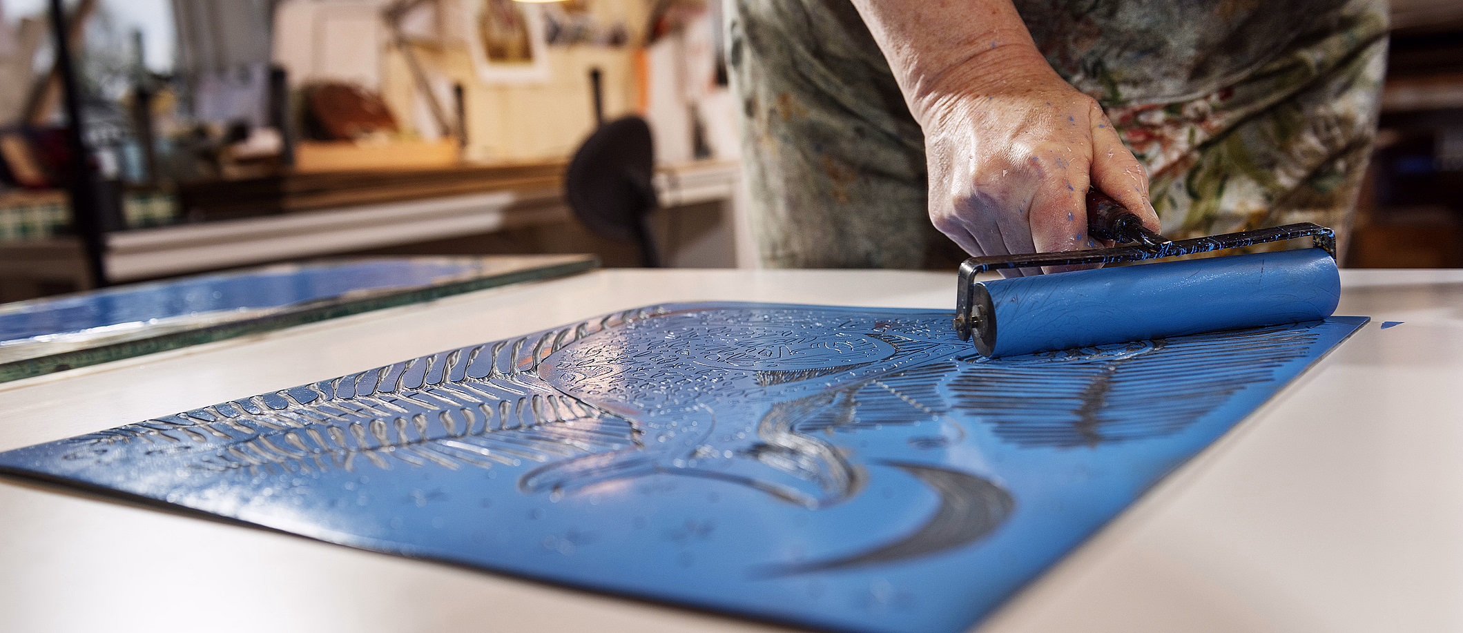 En person som påfører blått blekk på en utskåret linoleumplate med bladmønster ved bruk av en rulle i et kunststudio.
