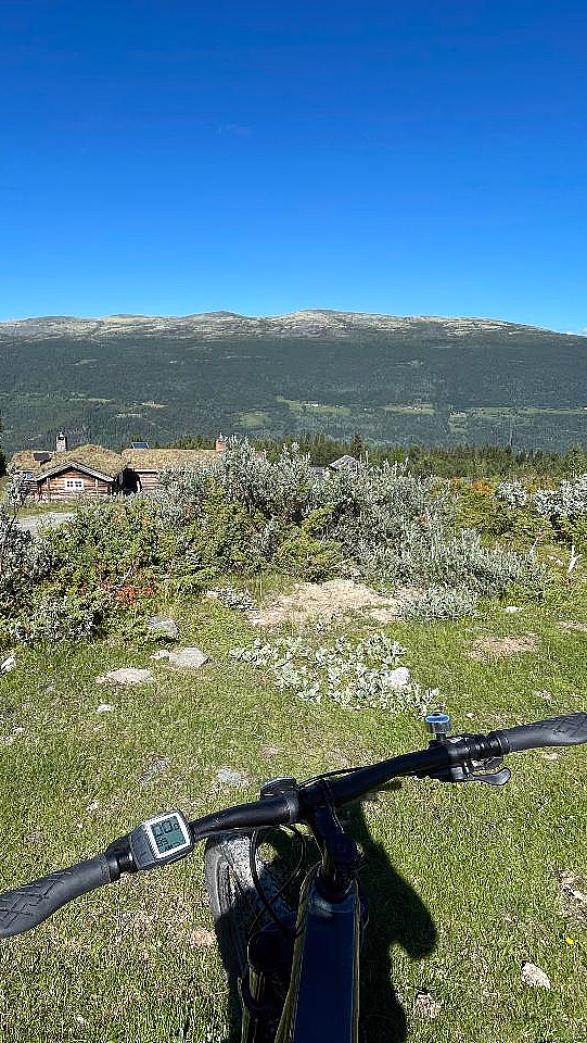 Sykkelstyre med digitalt display i forgrunnen på en gresskledd bakke, med små trehus med torvtak og en fjellkjede under en blå himmel i bakgrunnen.
