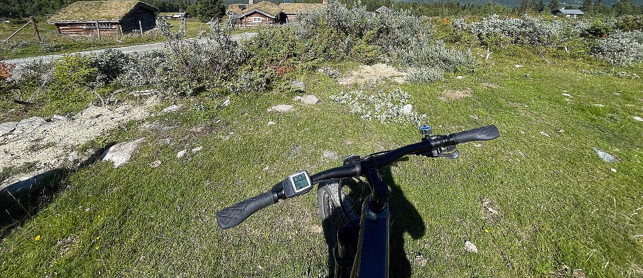 Sykkelstyre med digitalt display i forgrunnen på en gresskledd bakke, med små trehus med torvtak og en fjellkjede under en blå himmel i bakgrunnen.
