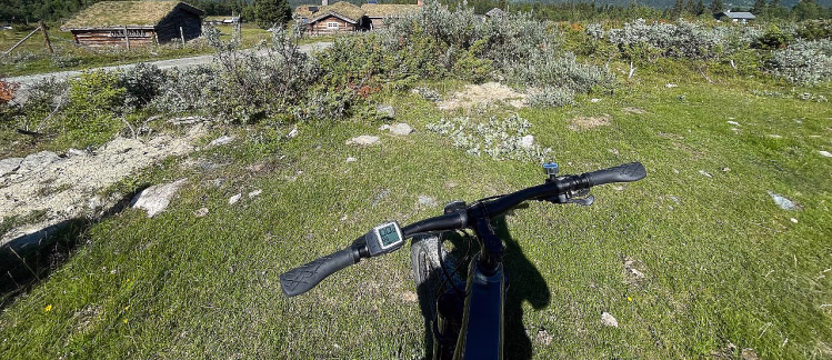 Sykkelstyre med digitalt display i forgrunnen på en gresskledd bakke, med små trehus med torvtak og en fjellkjede under en blå himmel i bakgrunnen.