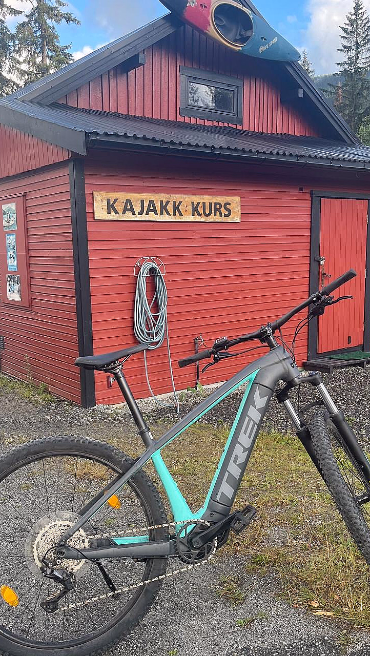 En rød trebygning med en kajakk på taket, en sykkel parkert foran og et skilt med teksten «KAJAKK KURS».