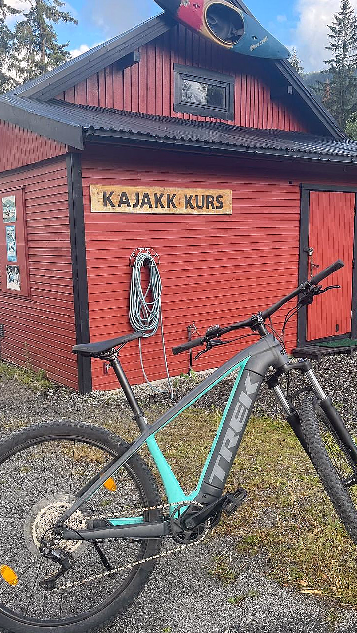 En rød trebygning med en kajakk på taket, en sykkel parkert foran og et skilt med teksten «KAJAKK KURS».