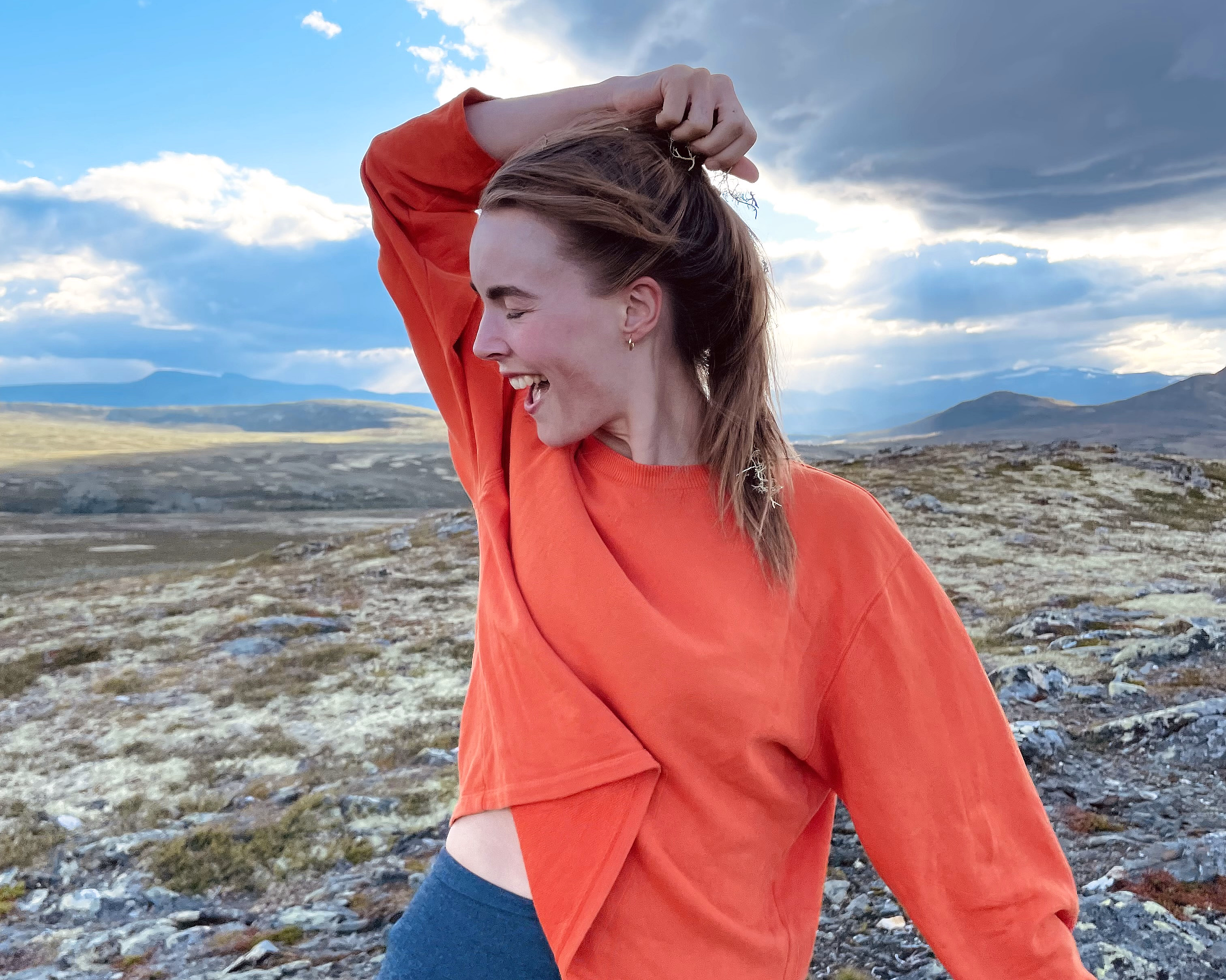 En smilende person i oransje langermet overdel og blå tights står på steinete terreng og holder i håret sitt, med fjell og skyer i bakgrunnen.