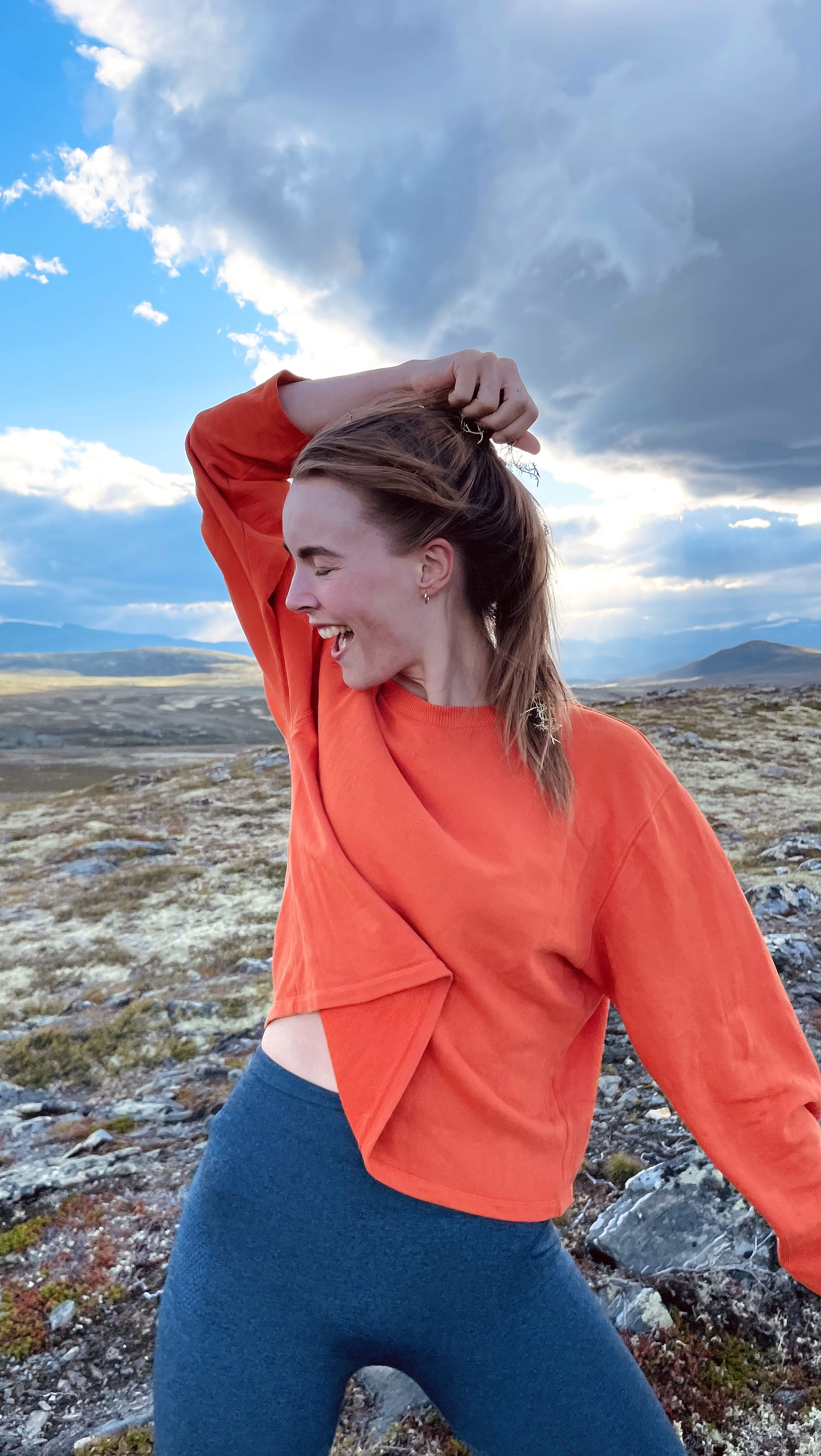 En smilende person i oransje langermet overdel og blå tights står på steinete terreng og holder i håret sitt, med fjell og skyer i bakgrunnen.
