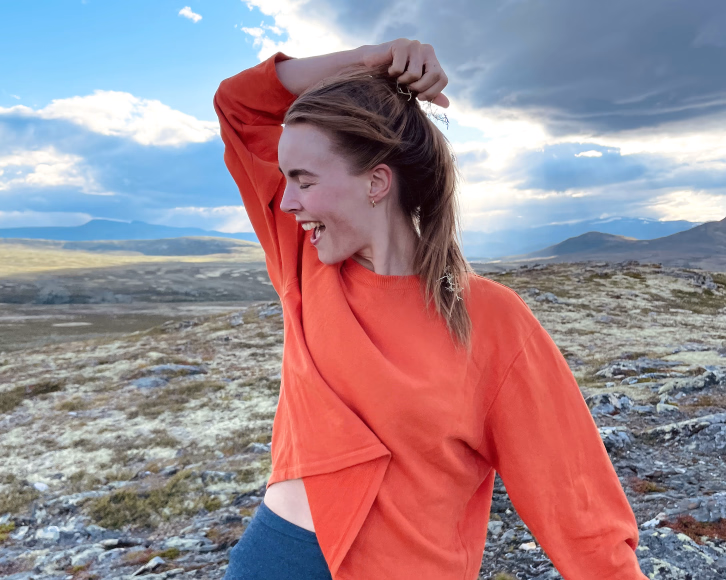 En smilende person i oransje langermet overdel og blå tights står på steinete terreng og holder i håret sitt, med fjell og skyer i bakgrunnen.