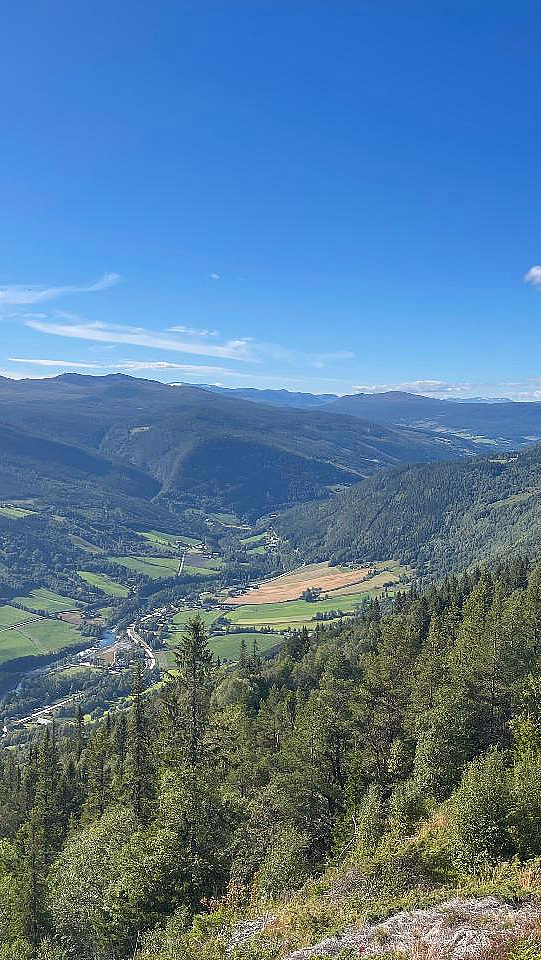 En frodig dal med en svingete elv, skogkledde åser og en liten hytte med torvtak under en blå himmel.