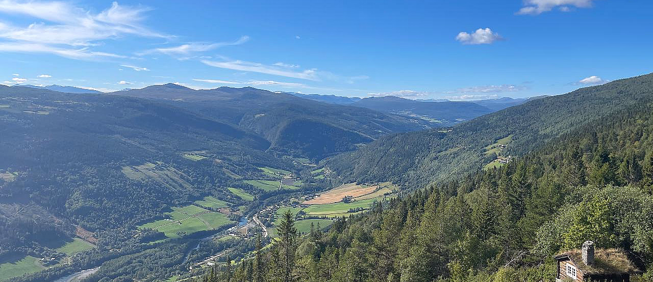 En frodig dal med en svingete elv, skogkledde åser og en liten hytte med torvtak under en blå himmel.