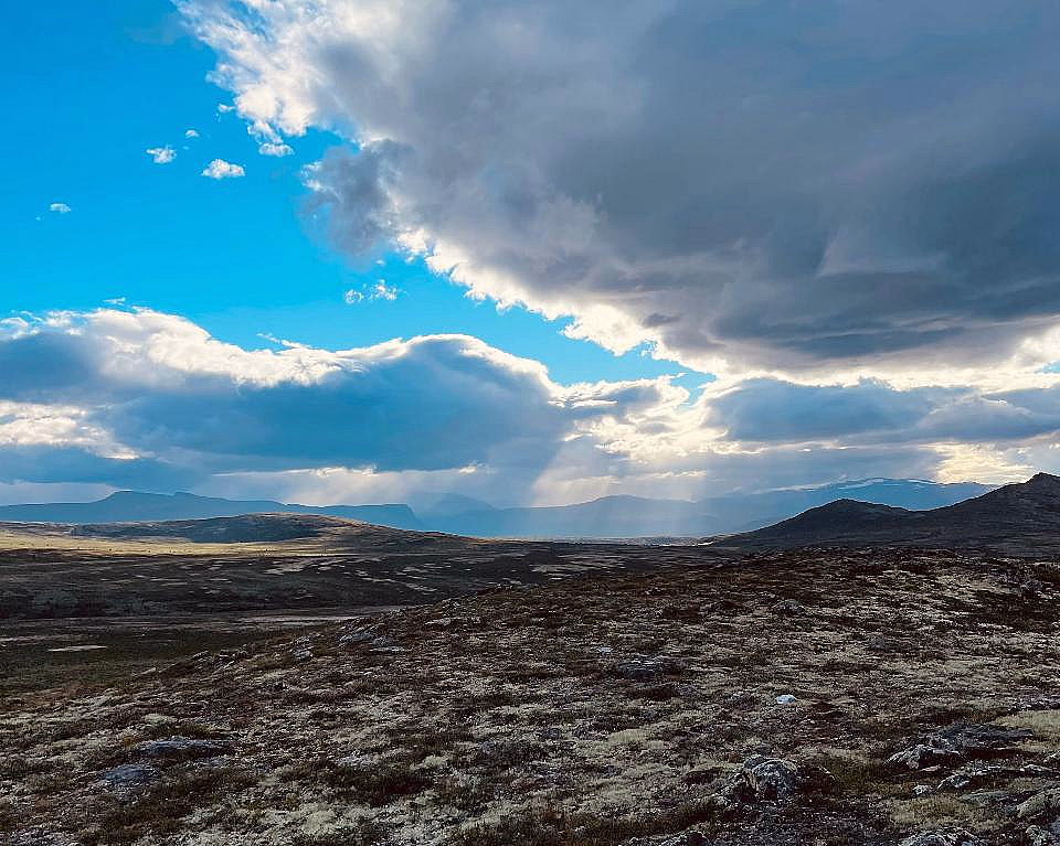 Et vidstrakt, steinete landskap med gress og en dramatisk himmel der solstråler bryter gjennom mørke skyer over fjerne fjell.