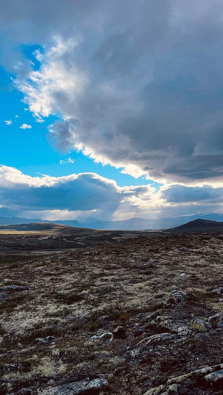 Et vidstrakt, steinete landskap med gress og en dramatisk himmel der solstråler bryter gjennom mørke skyer over fjerne fjell.