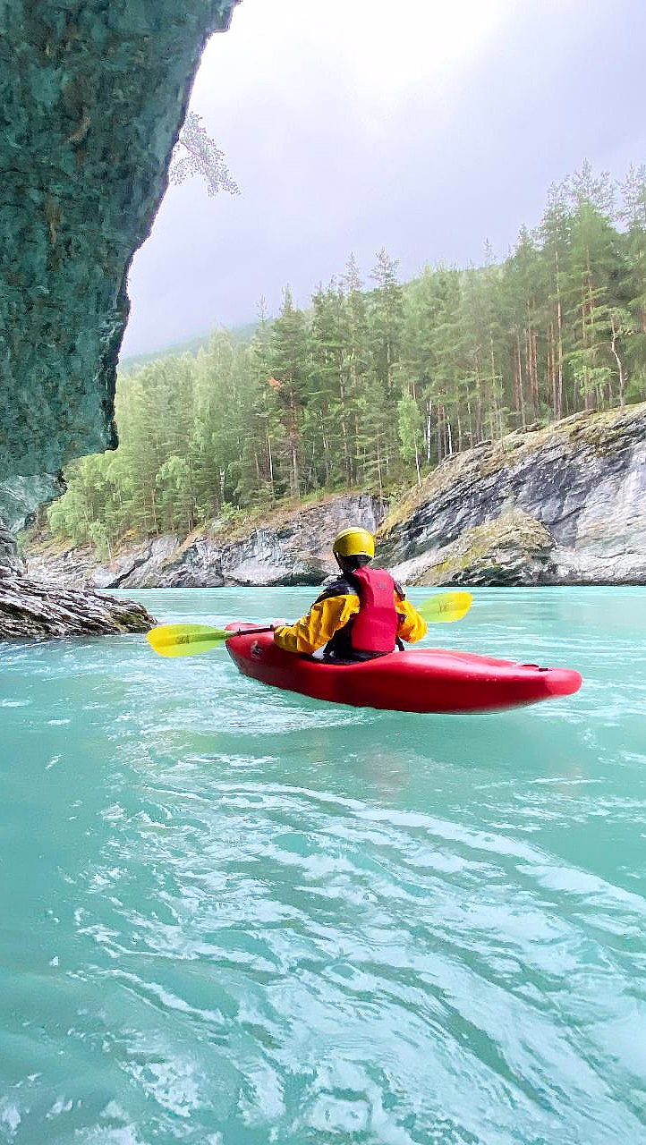 En person i rød kajakk med gul hjelm og rød redningsvest padler på turkist vann under et klippefremspring.