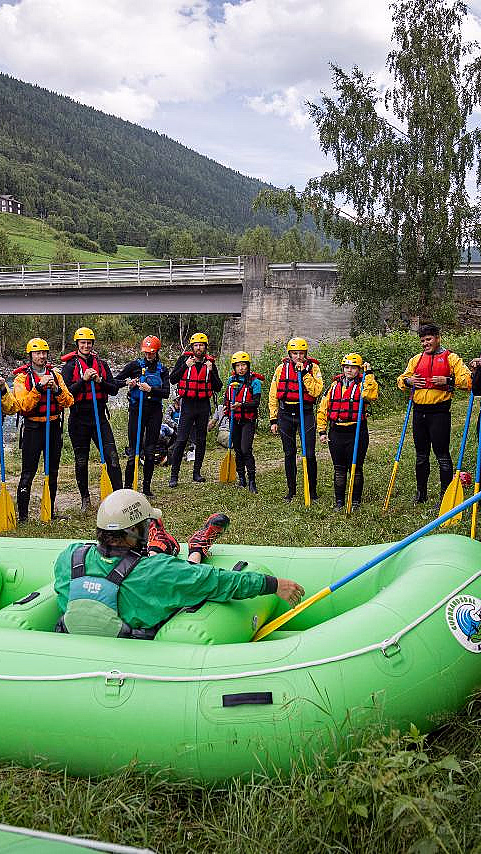 En gruppe mennesker i gule hjelmer og røde redningsvester står med padleårer ved en grønn flåte, med en elv, en bro og en ås i bakgrunnen.