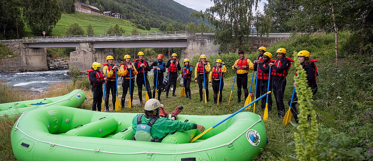 En gruppe mennesker i gule hjelmer og røde redningsvester står med padleårer ved en grønn flåte, med en elv, en bro og en ås i bakgrunnen.