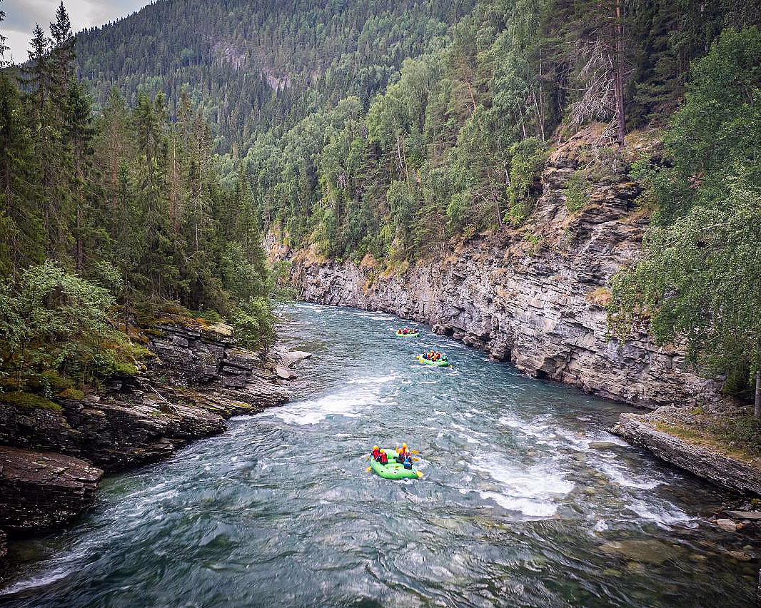 Rafting i grønne gummibåter på en elv som renner mellom bratte klipper og tett skog.