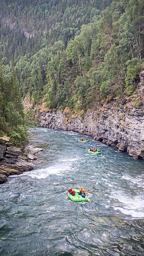 Rafting i grønne gummibåter på en elv som renner mellom bratte klipper og tett skog.