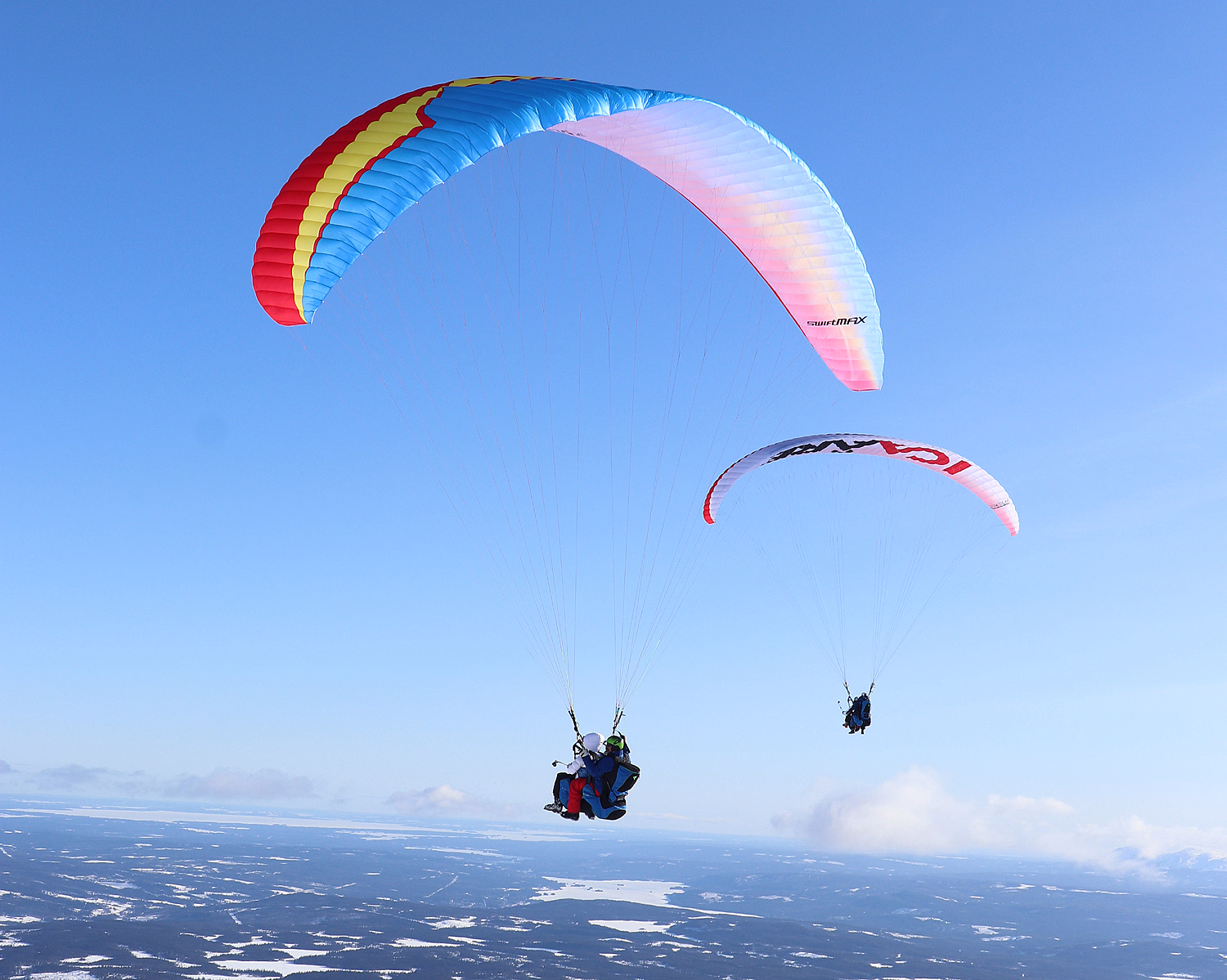 To paraglidere flyr over et snødekt landskap under en klar blå himmel. Den ene vingen er blå med rød og gul kant, mens den andre er hvit med rød og svart tekst.