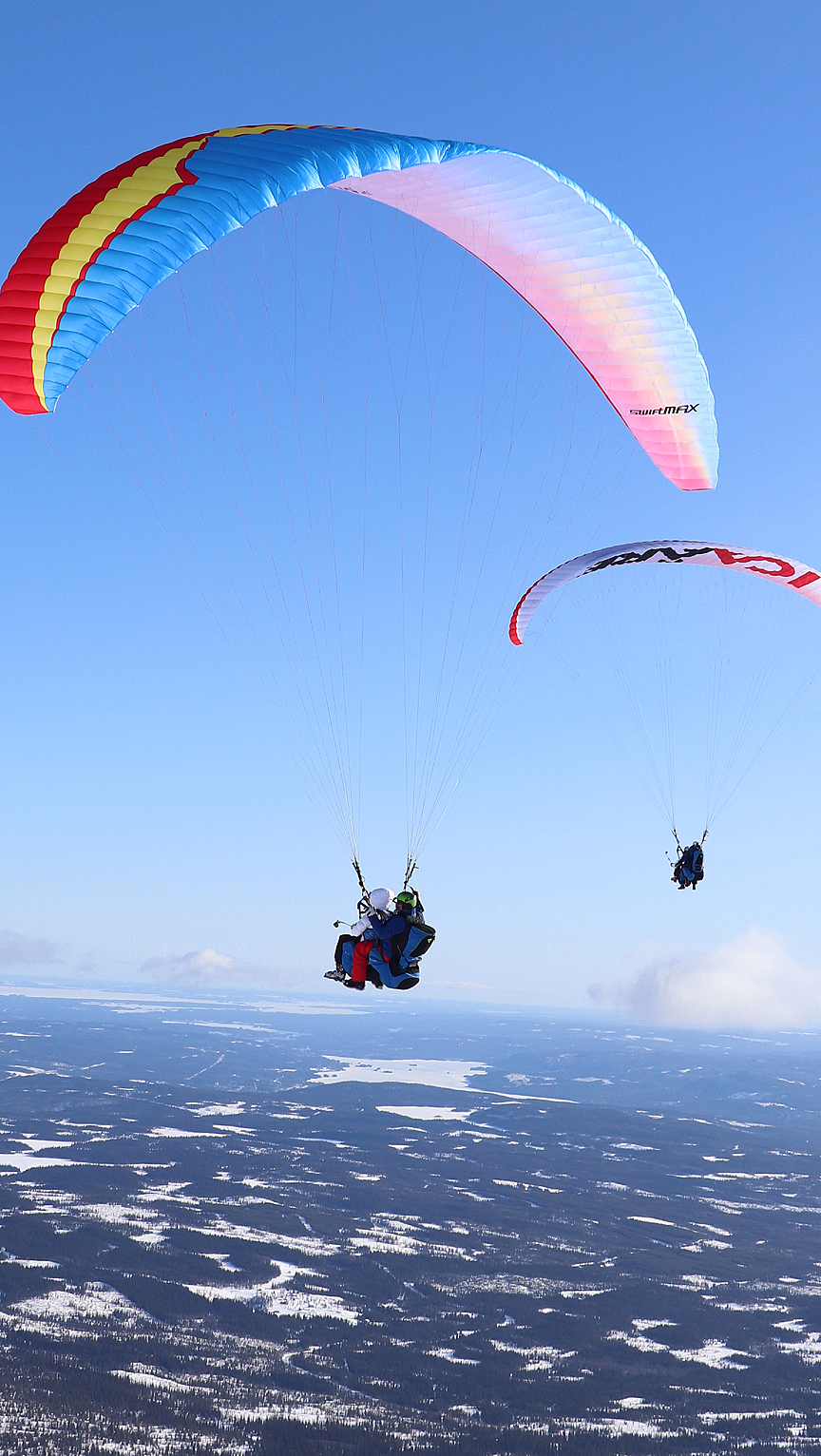To paraglidere flyr over et snødekt landskap under en klar blå himmel. Den ene vingen er blå med rød og gul kant, mens den andre er hvit med rød og svart tekst.